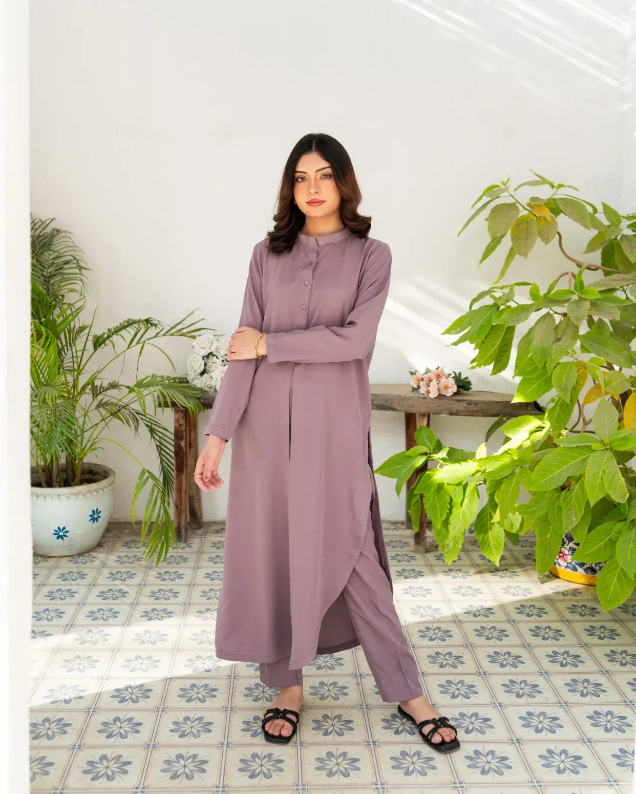 Woman in a long lavender color solid dress standing in a room with plants and a tiled floor.