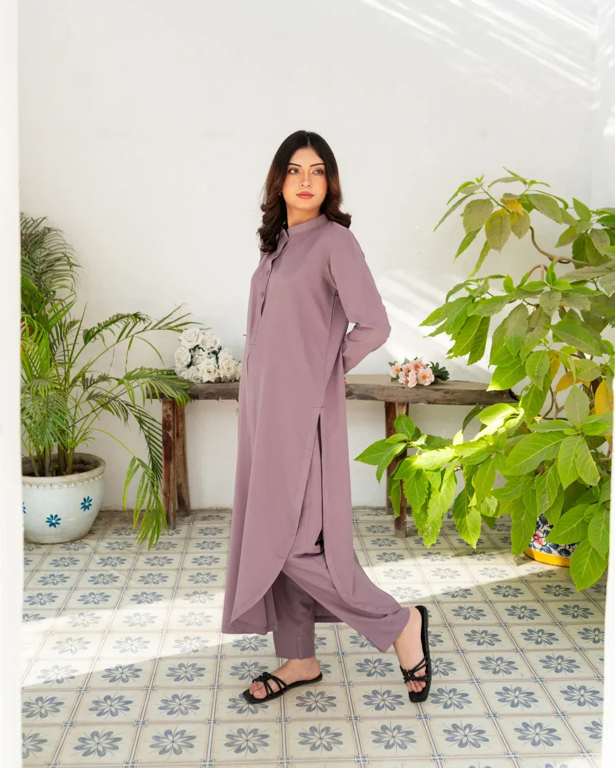 Woman in a lavender outfit standing in a room with plants and a tiled floor.