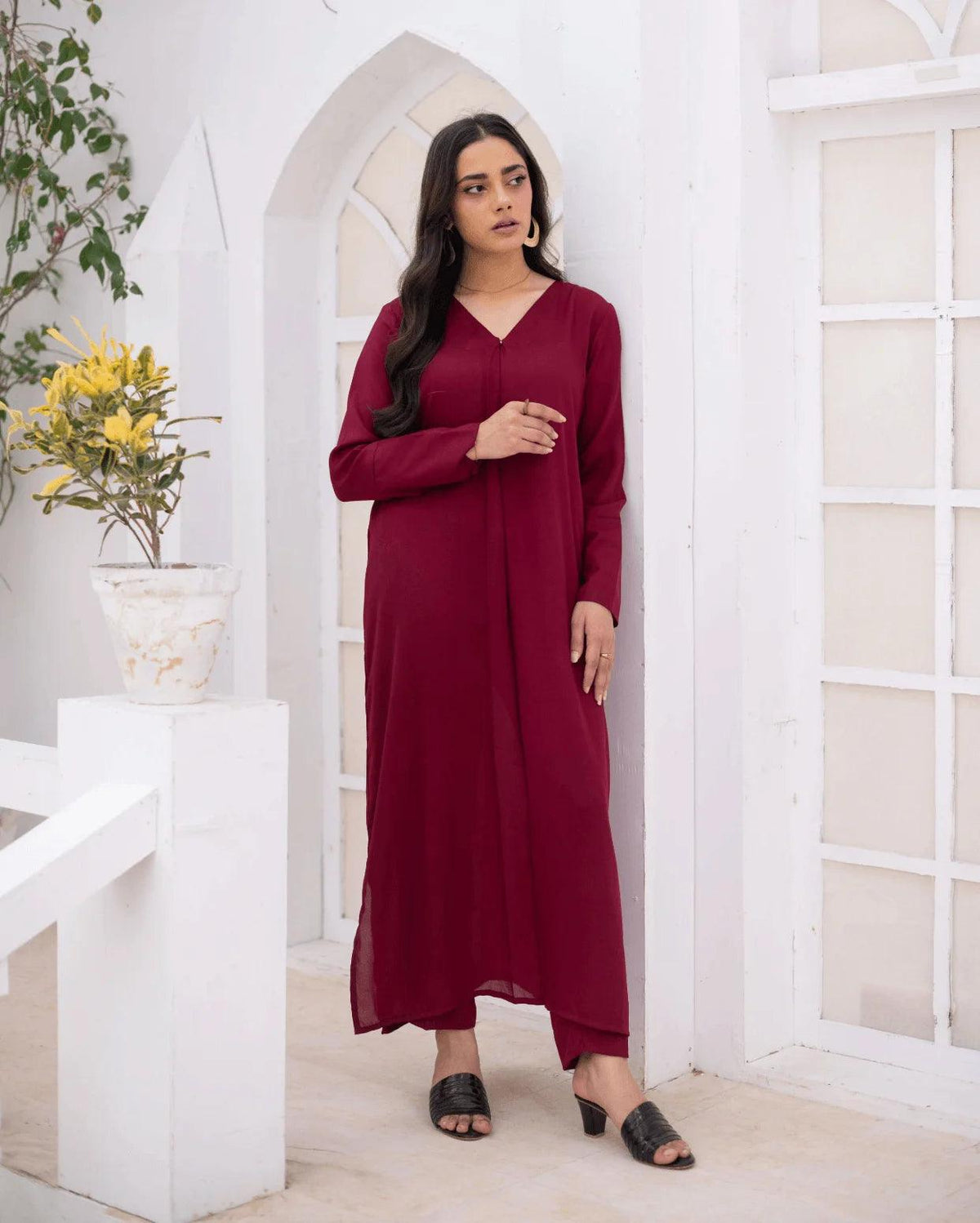A woman wearing a long-sleeved, V-neck co ord set in margenta, standing in a white arched corridor with a potted plant on a white pedestal.