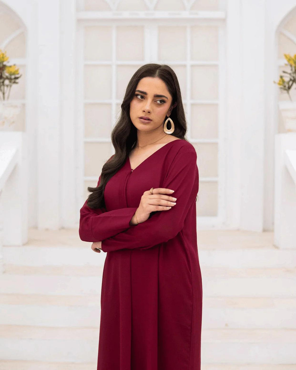 A woman with long dark hair stands in a white architectural setting wearing a long-sleeved magenta co ord set and large gold hoop earrings.