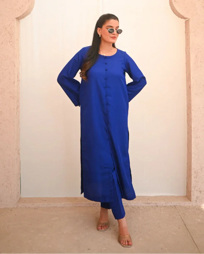 A woman standing and posing in a blue two-piece rawsilk dress with a shirt and straight trousers.
