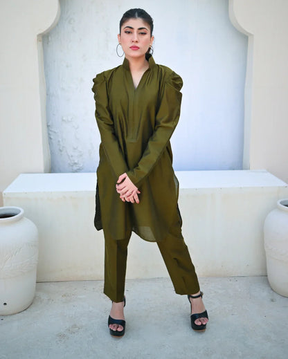 Woman in a green outfit with a white wall and decorative elements in the background.