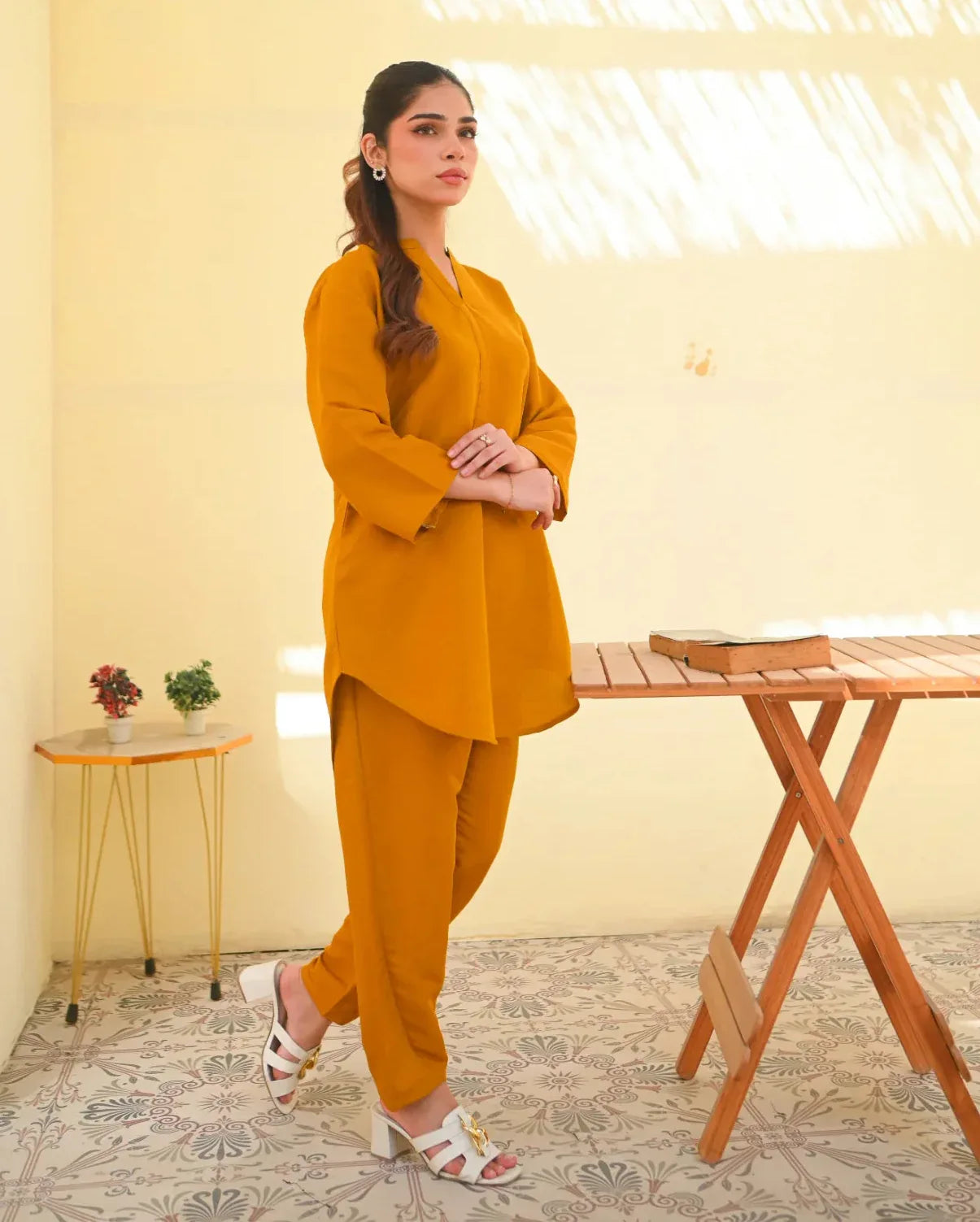 A woman wearing a mustard yellow two-piece dress with long sleeves, consisting of a shirt and trousers, standing in a sunny area next to a wooden bench.