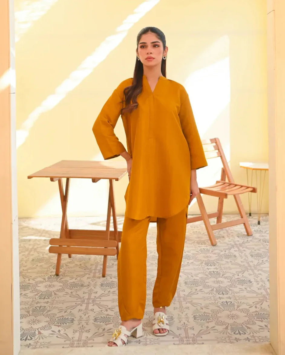 A woman wearing a mustard yellow two-piece dress with long sleeves, consisting of a shirt and trousers, standing in a sunny area next to a wooden bench.