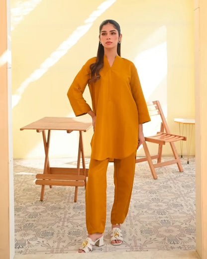 A woman wearing a mustard yellow two-piece dress with long sleeves, consisting of a shirt and trousers, standing in a sunny area next to a wooden bench.