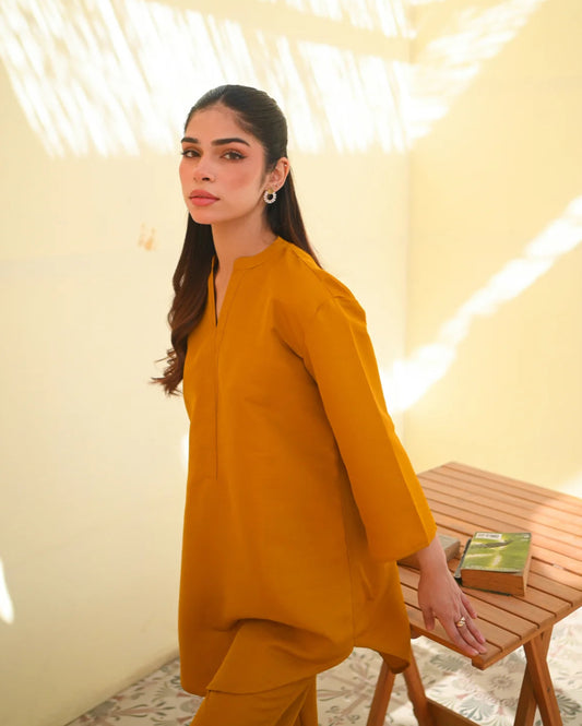 A woman wearing a mustard yellow two-piece dress with long sleeves, consisting of a shirt and trousers, standing in a sunny area next to a wooden bench.