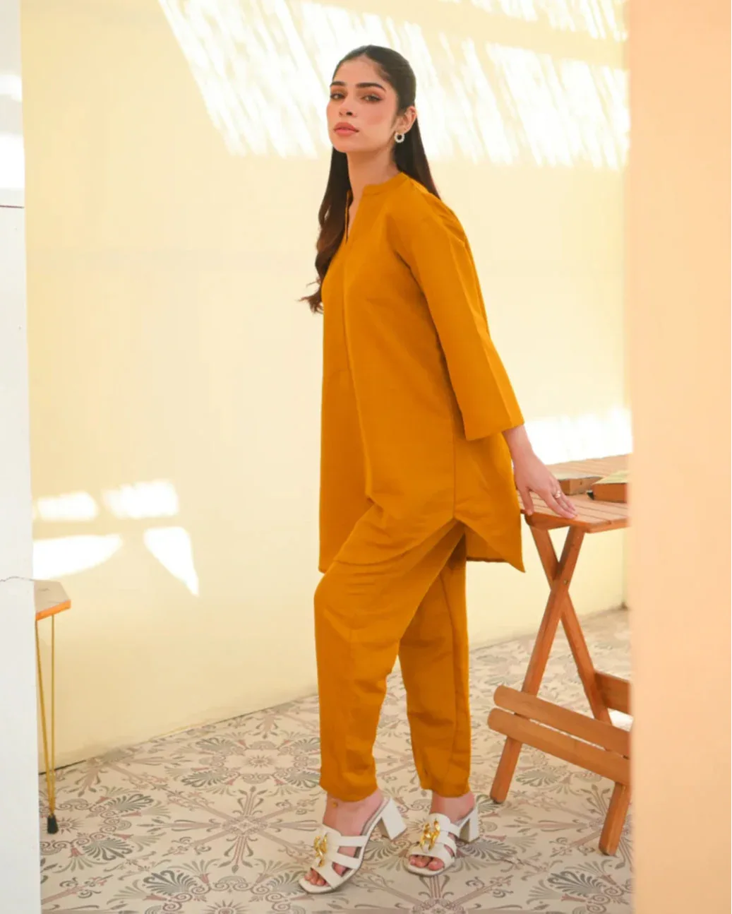 A woman wearing a mustard yellow two-piece dress with long sleeves, consisting of a shirt and trousers, standing in a sunny area next to a wooden bench.