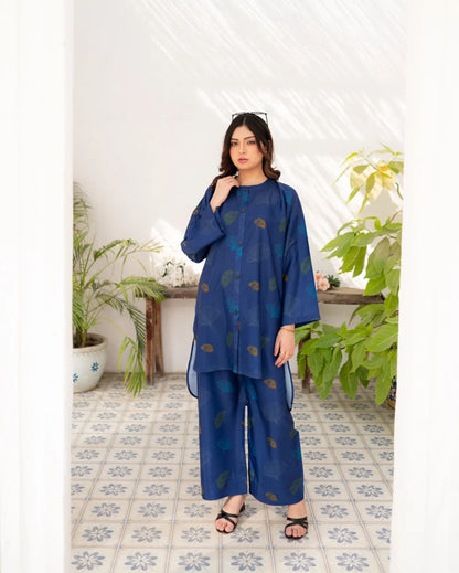 Woman wearing a navy blue outfit with a pattern in a room with plants and a white wall.