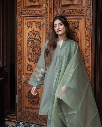 A woman in an olive green stitched dress stands confidently against an ornately carved wooden door, conveying elegance and cultural richness.