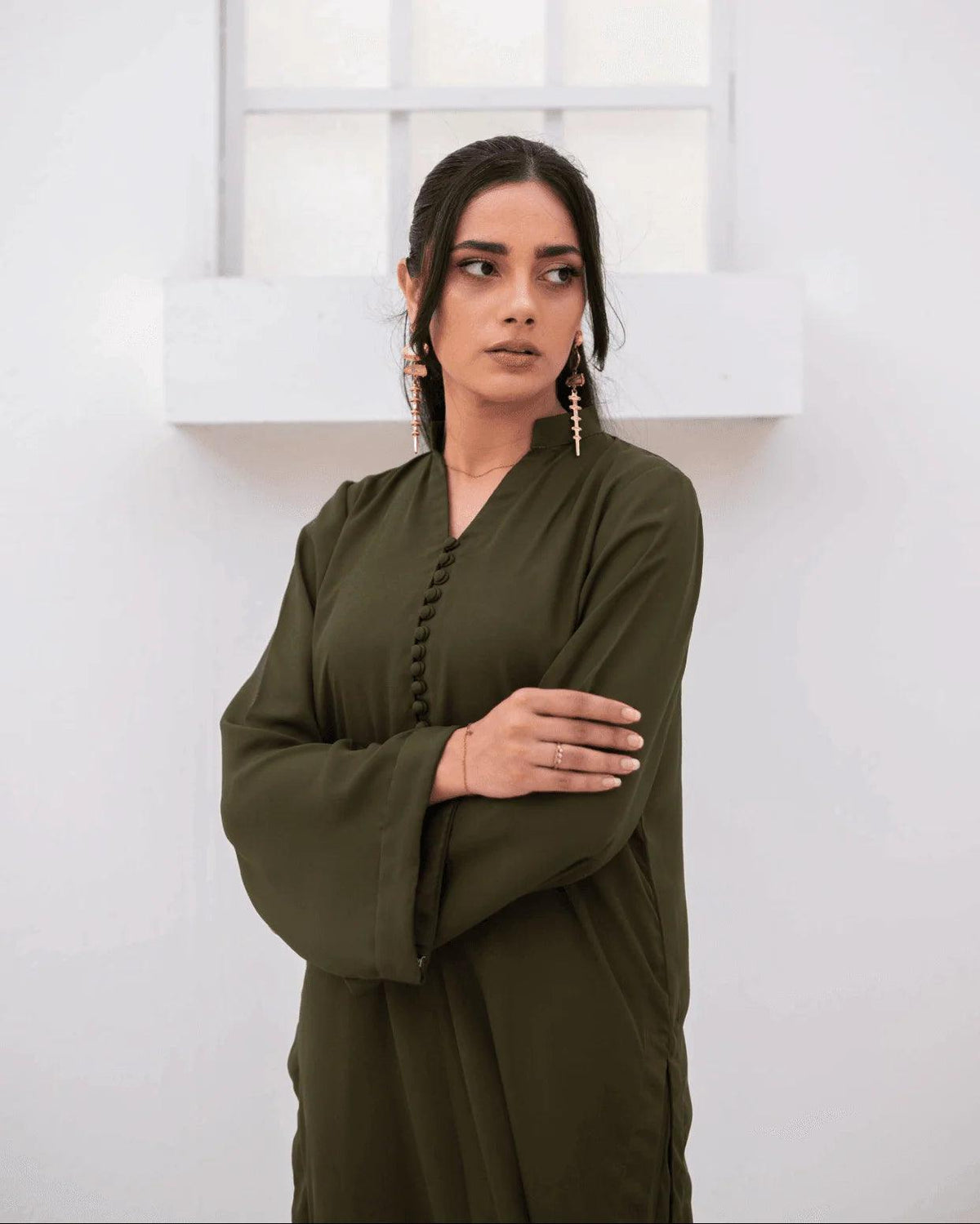 A woman wearing a long-sleeved olive green co ord set with buttoned V-neckline, standing against a white wall with a window frame.