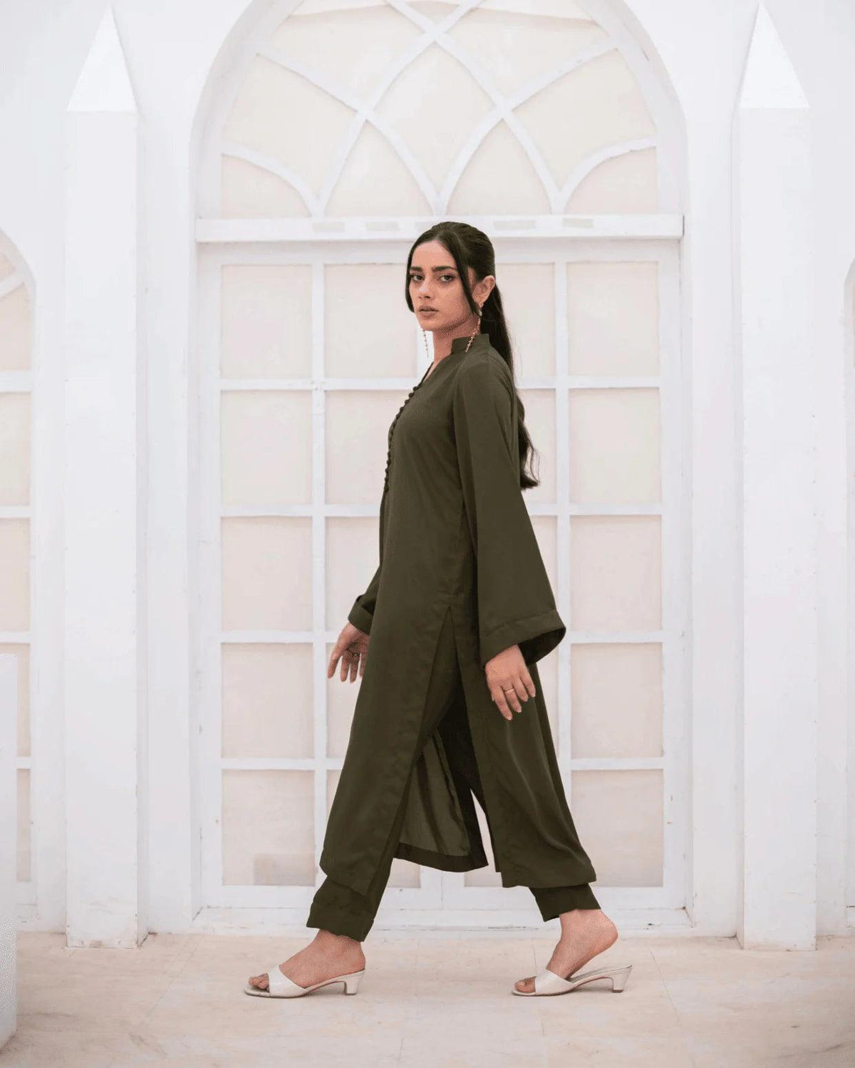 A woman wearing an olive green co ord set walks in profile in front of a white arched window frame.
