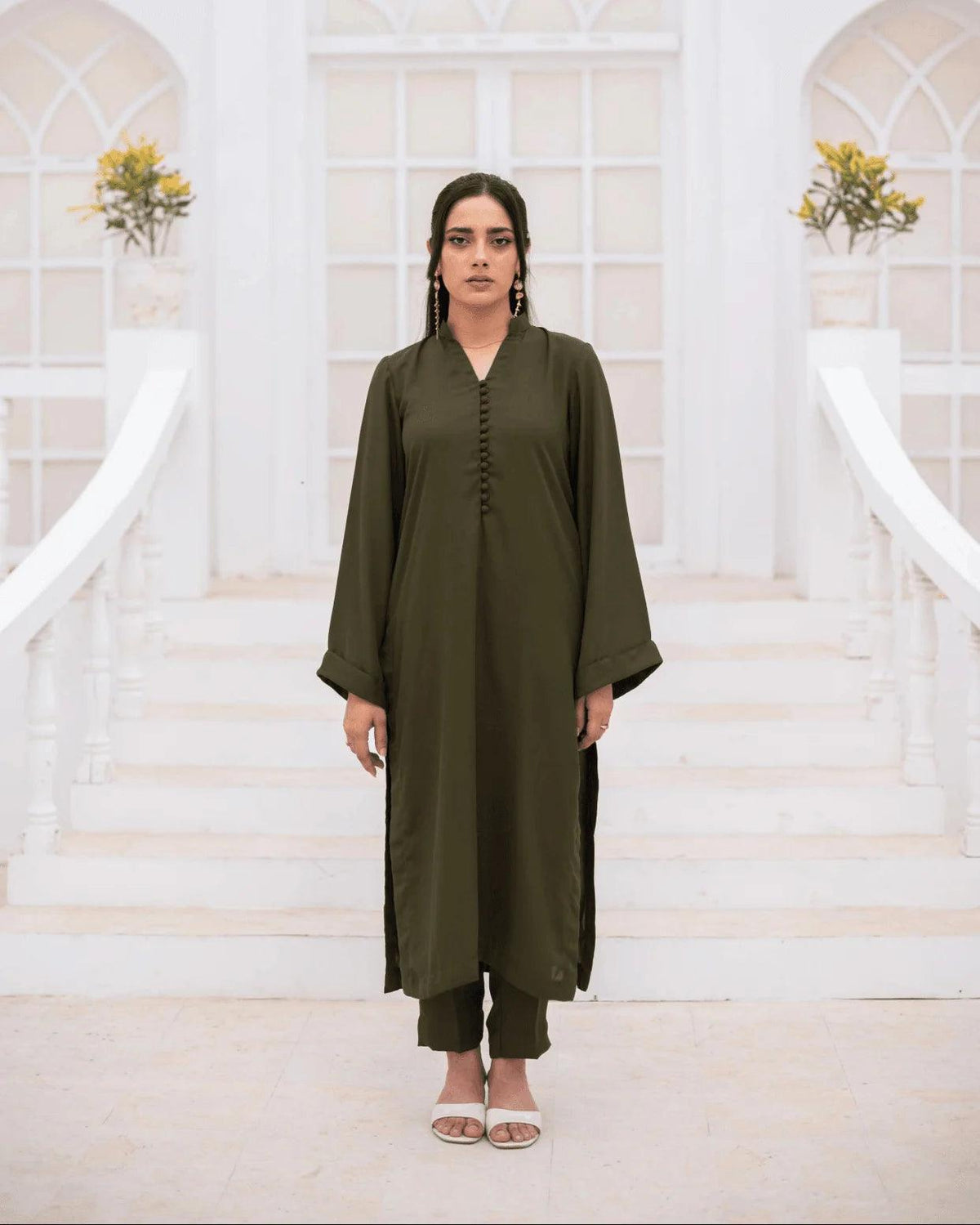 A woman stands centered on white stairs wearing an olive green long kurta-style co ord set. The setting has white arched windows and small flower arrangements in the background.