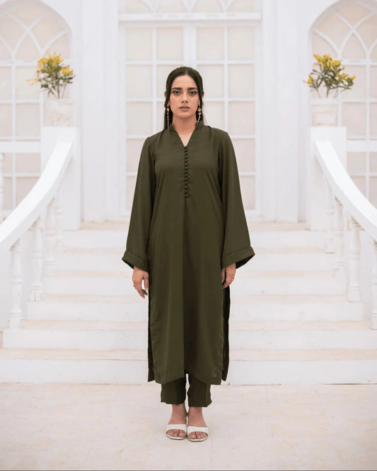 A woman stands centered on white stairs wearing an olive green long kurta-style co ord set. The setting has white arched windows and small flower arrangements in the background.