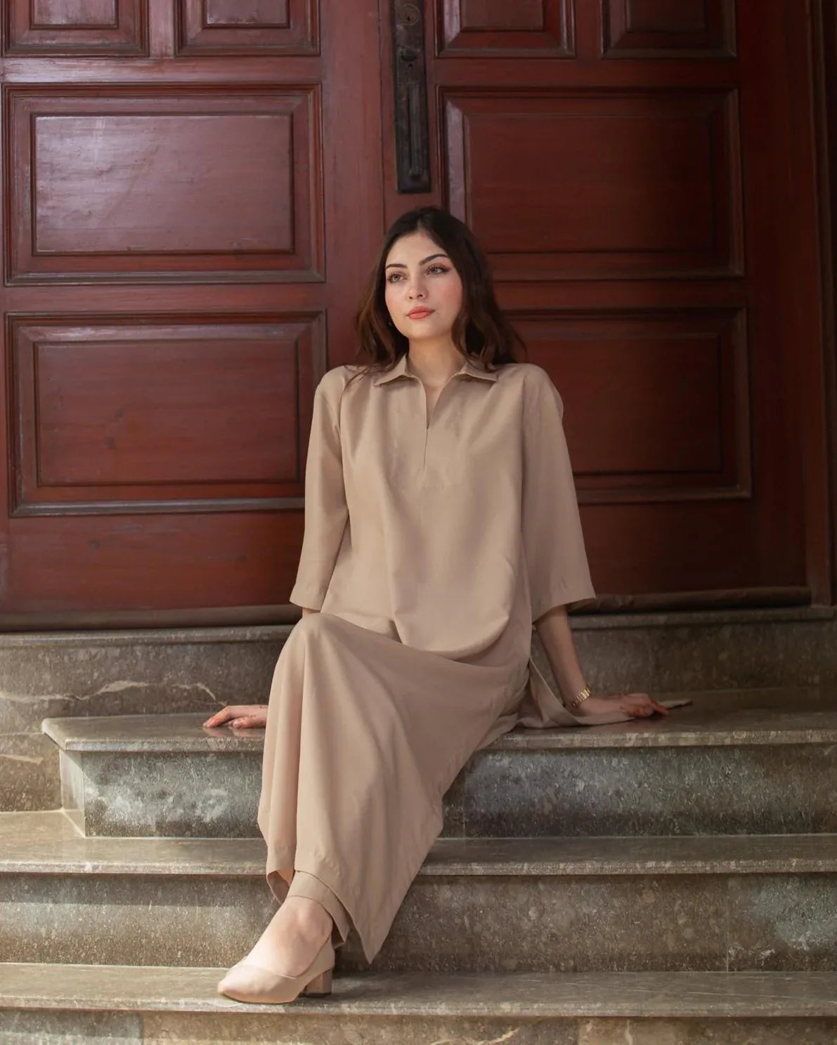 A woman in a phone outfit sitting in front of large wooden doors with a thoughtful expression. 