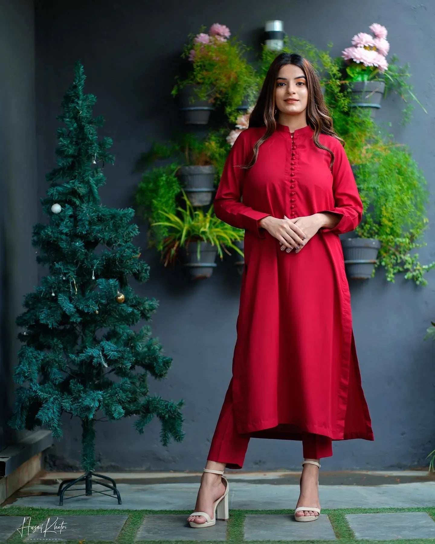 Woman in a red dress stands confidently near a small decorated tree, with a backdrop of green plants on a gray wall, conveying elegance.
