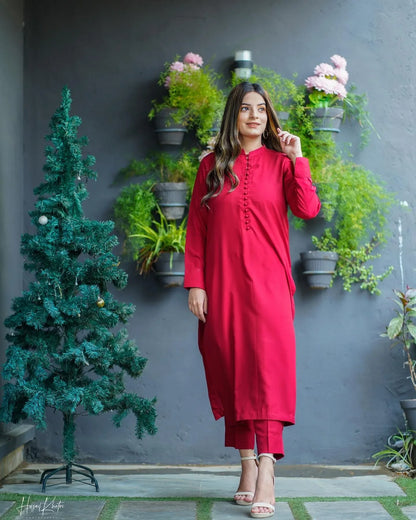 Woman in a red dress stands confidently near a small decorated tree, with a backdrop of green plants on a gray wall, conveying elegance.