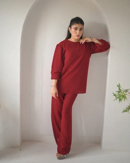 A woman stands in an arched alcove wearing a loose-fitting, long-sleeved red top and matching red trousers, with minimal jewelry and light-colored heels.