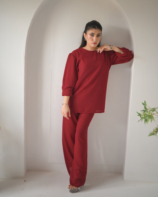 A woman stands in an arched alcove wearing a loose-fitting, long-sleeved red top and matching red trousers, with minimal jewelry and light-colored heels.