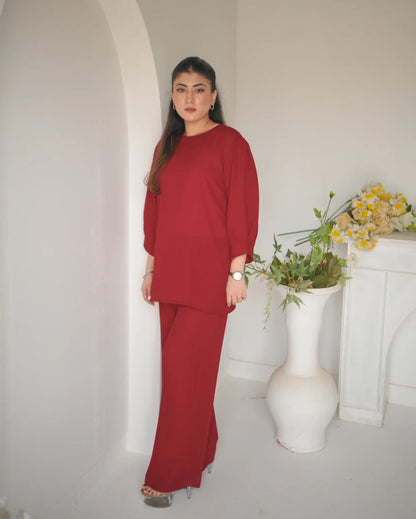 A woman standing and wearing a loose-fitting, long-sleeved red top and matching red trousers, with minimal jewelry and light-colored heels.