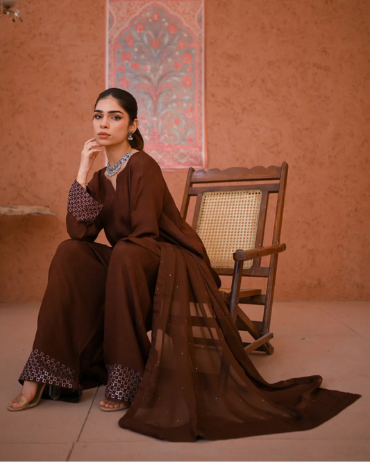 A woman wearing a brown three-piece rawsilk dress which includes a shirt and farshi shalwar along with a chiffon dupatta. The dress is embellished with stone details.