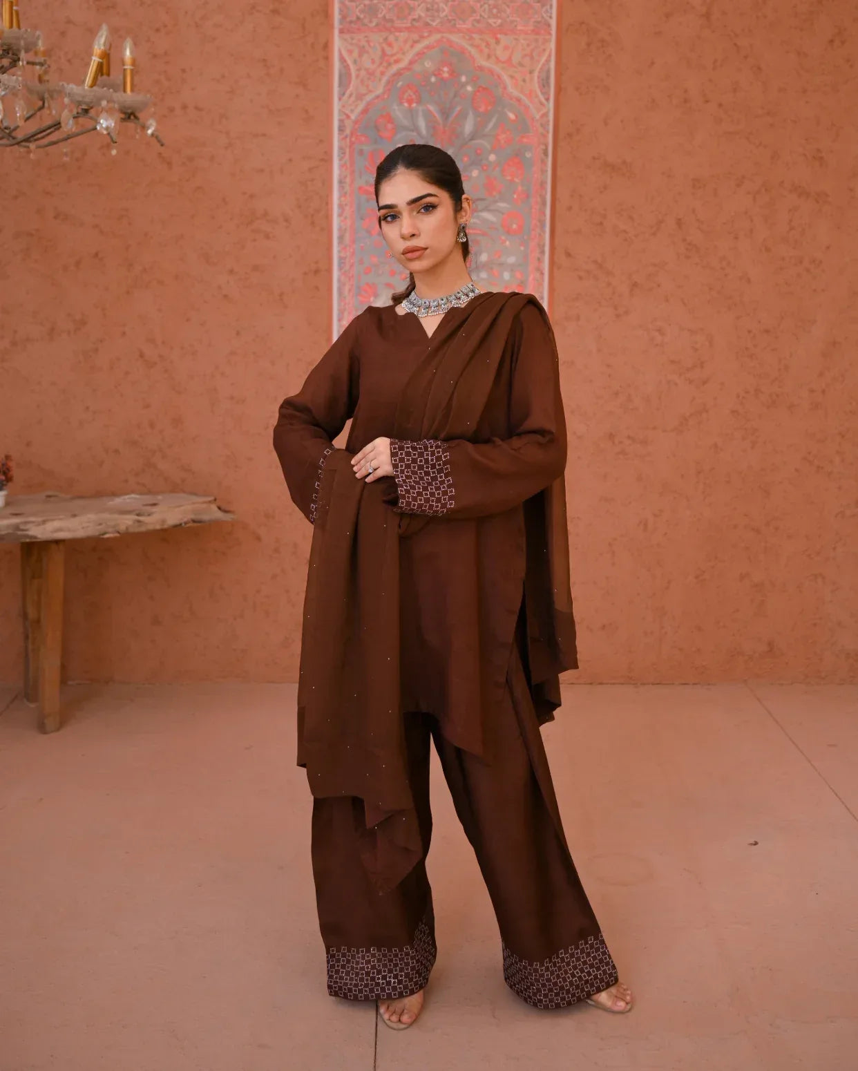 A woman wearing a brown three-piece rawsilk dress which includes a farshi shalwar kameez along with a chiffon dupatta. The dress is embellished with stone details