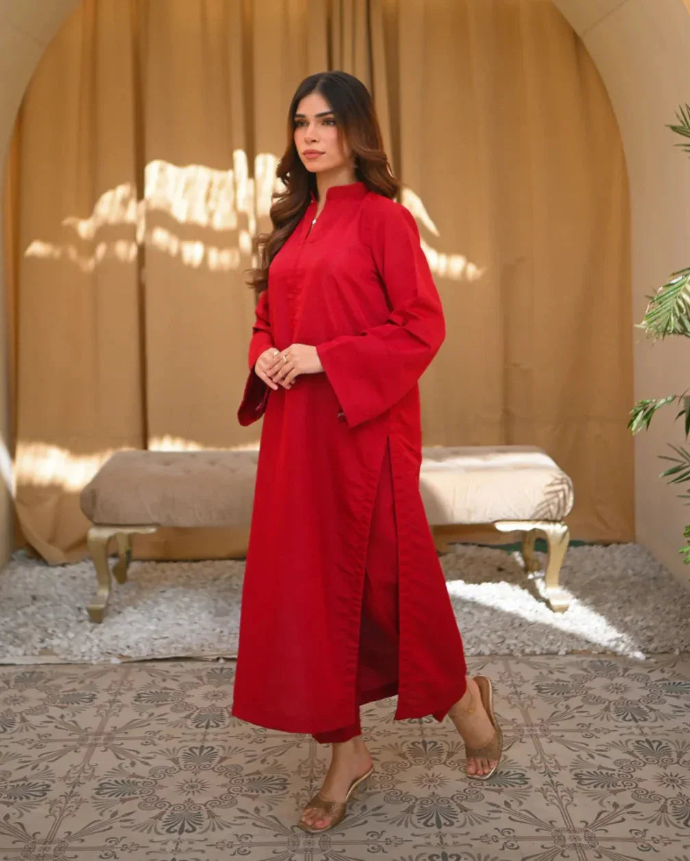 A woman standing in a sunny room wearing a red two-piece rawsilk dress with a long shirt and straight trousers, posing next to a beige bench and a green plant.