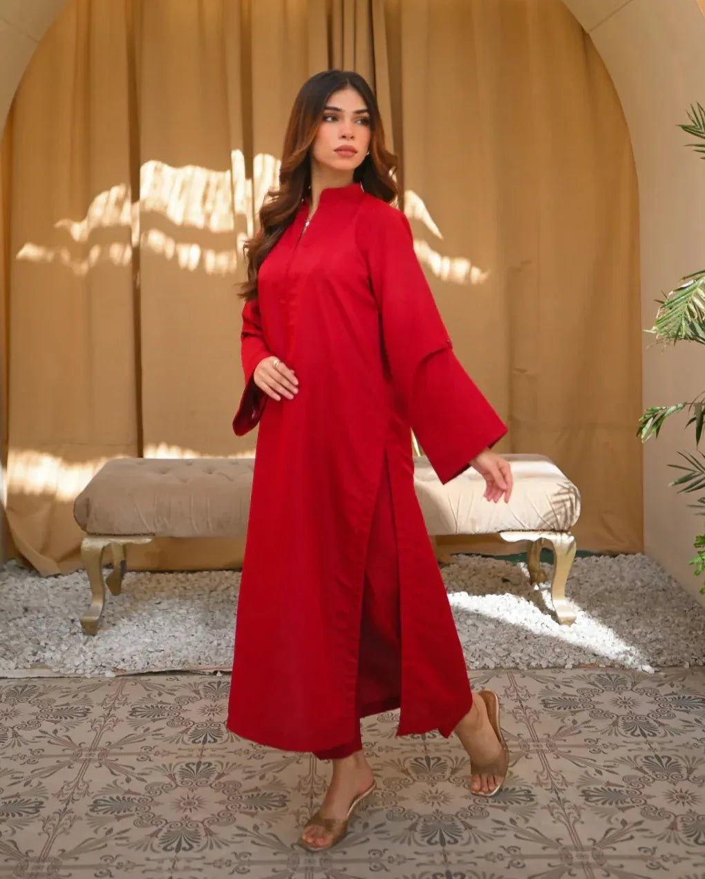 A woman standing in a sunny room wearing a red two-piece rawsilk dress with a long shirt and straight trousers, posing next to a beige bench and a green plant.