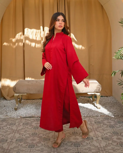 A woman standing in a sunny room wearing a red two-piece rawsilk dress with a long shirt and straight trousers, posing next to a beige bench and a green plant.
