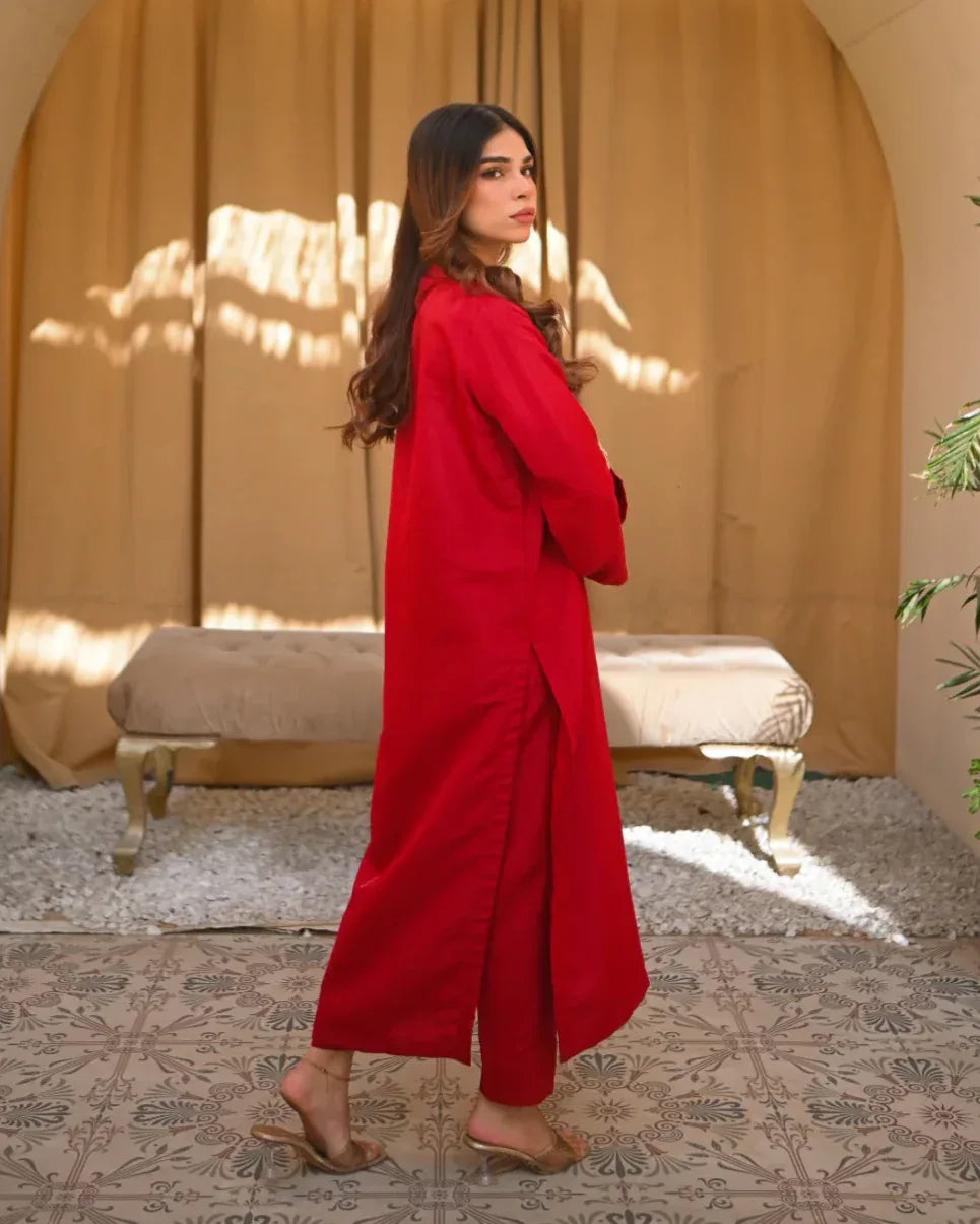 A woman standing in a sunny room wearing a red two-piece rawsilk dress with a long shirt and straight trousers, posing next to a beige bench and a green plant.