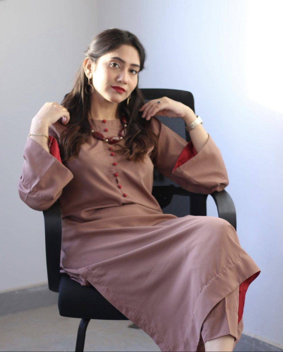 A woman in a brown dress with red accents sits casually on a black chair.