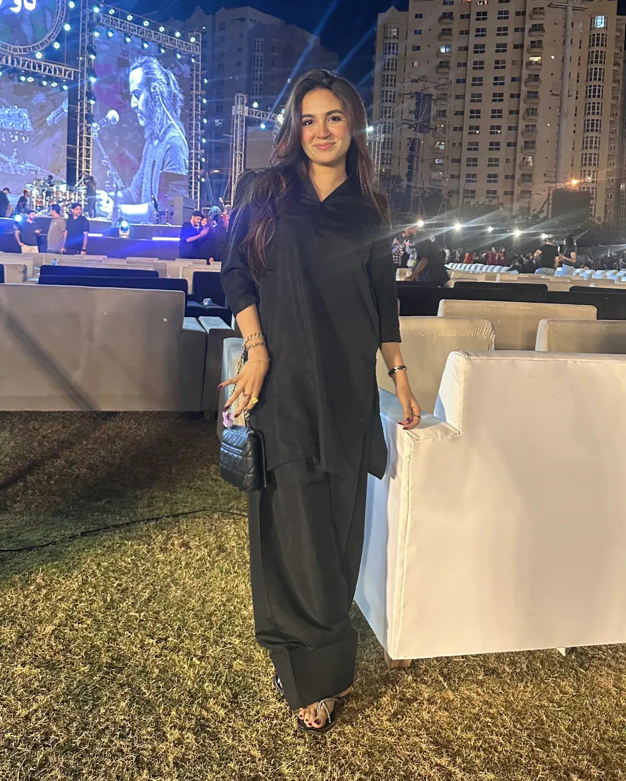 Woman in black farshi shalwar kameez standing in front of a large screen displaying a person, with buildings and event setup in the background.