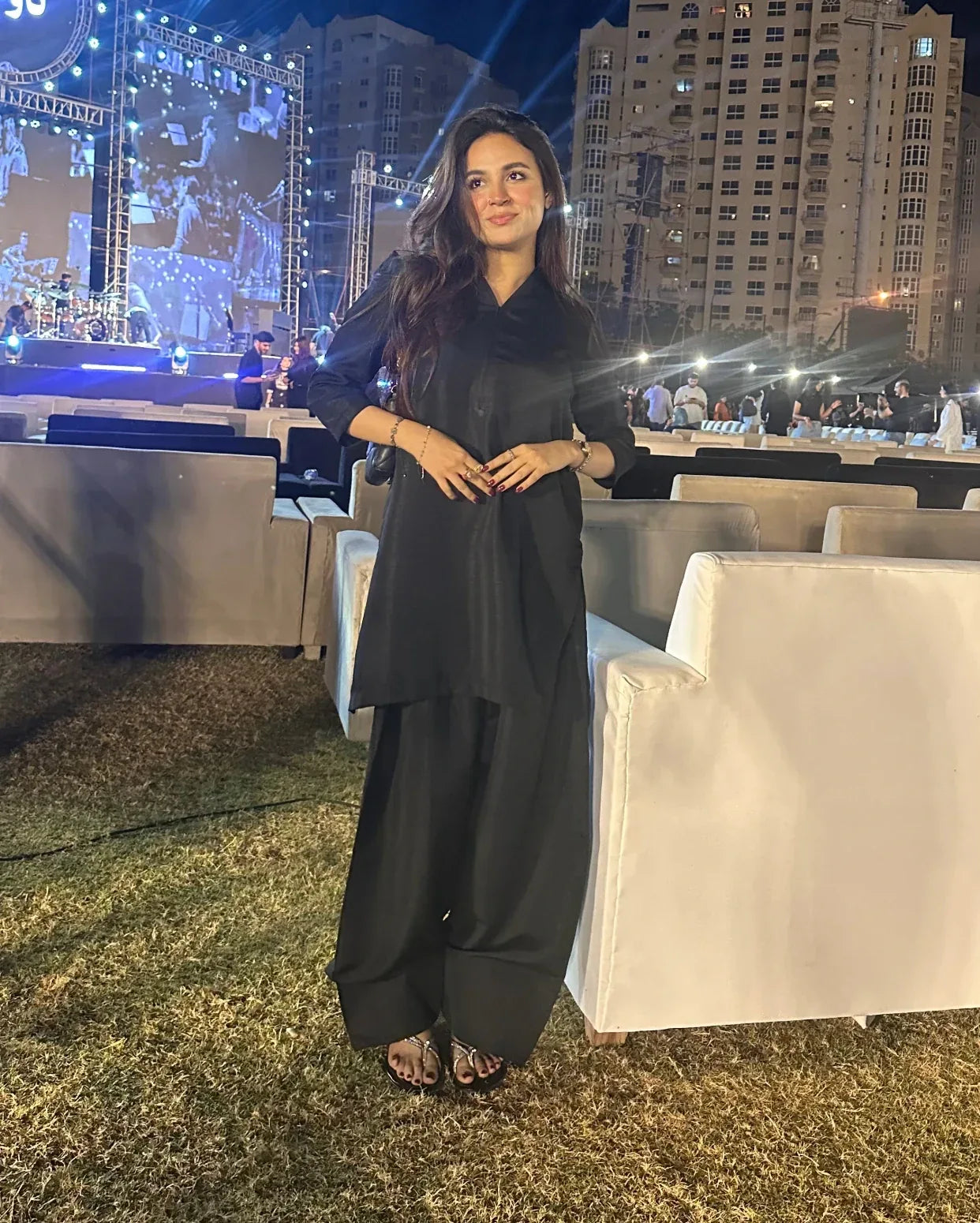 Woman in a black farshi shalwar kameez standing outdoors at an event with buildings and people in the background.