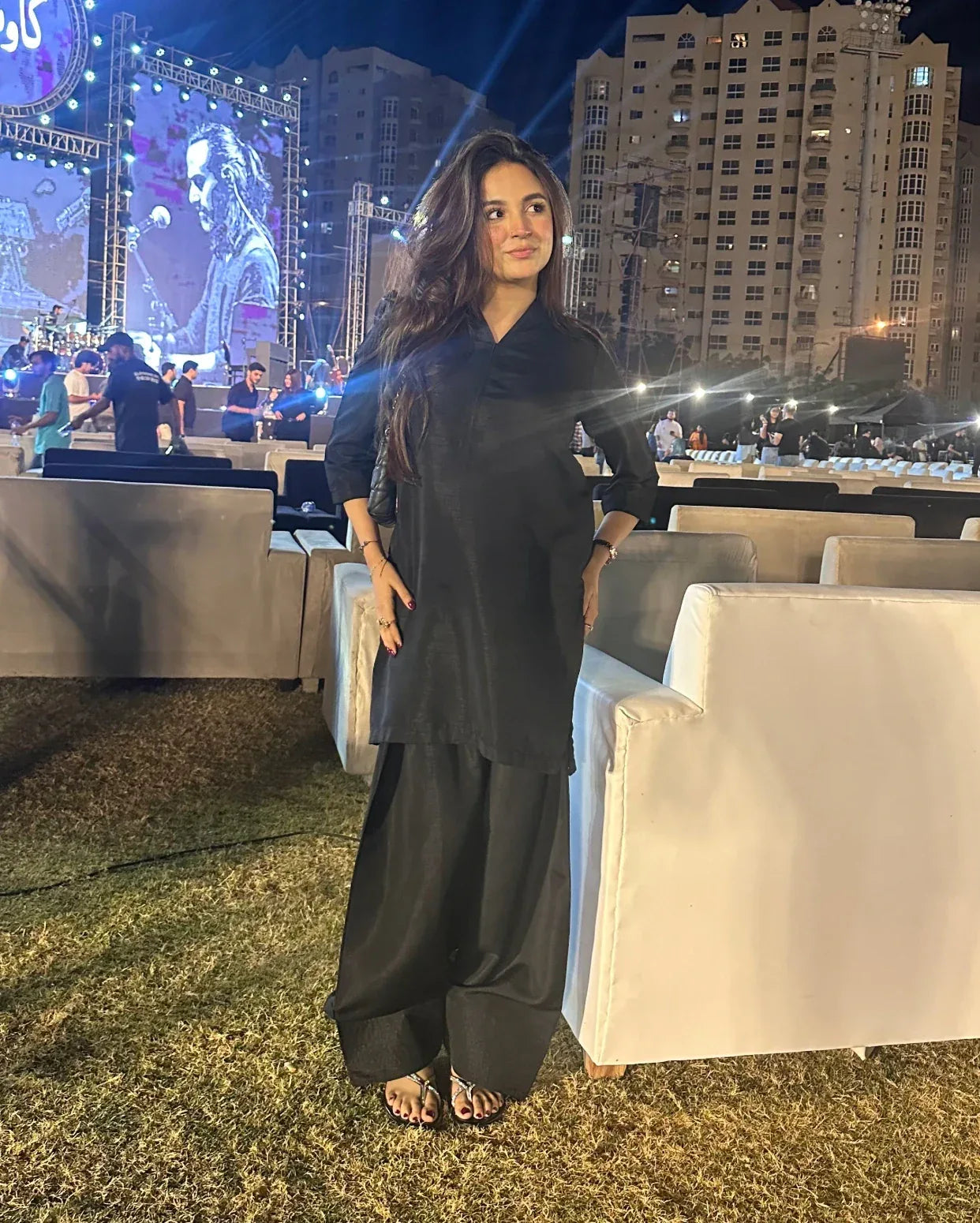 Woman in a black farshi shalwar kameez standing outdoors at an event with large screens and buildings in the background.