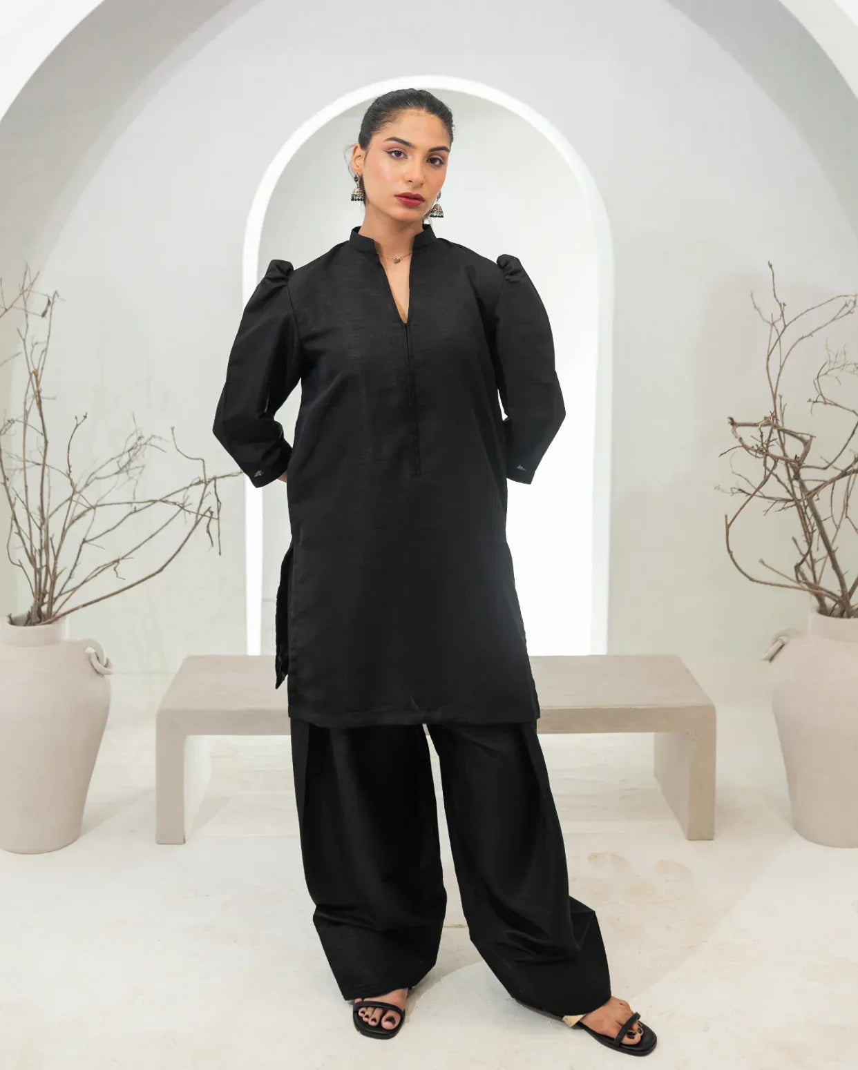 Woman wearing a black traditional two piece farshi shalwar kameez in a minimalistic setting with white arches and potted plants.