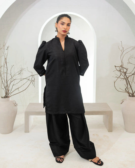 Woman wearing a black traditional two piece kameez and farshi shawlar outfit in a minimalistic setting with white arches and potted plants.