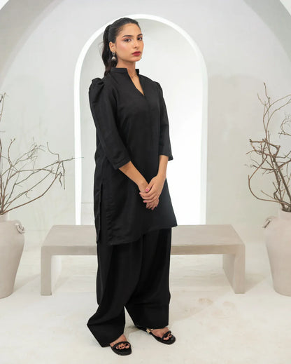 Woman wearing a black two piece farshi shalwar kameez standing in a minimalistic white room with plants.