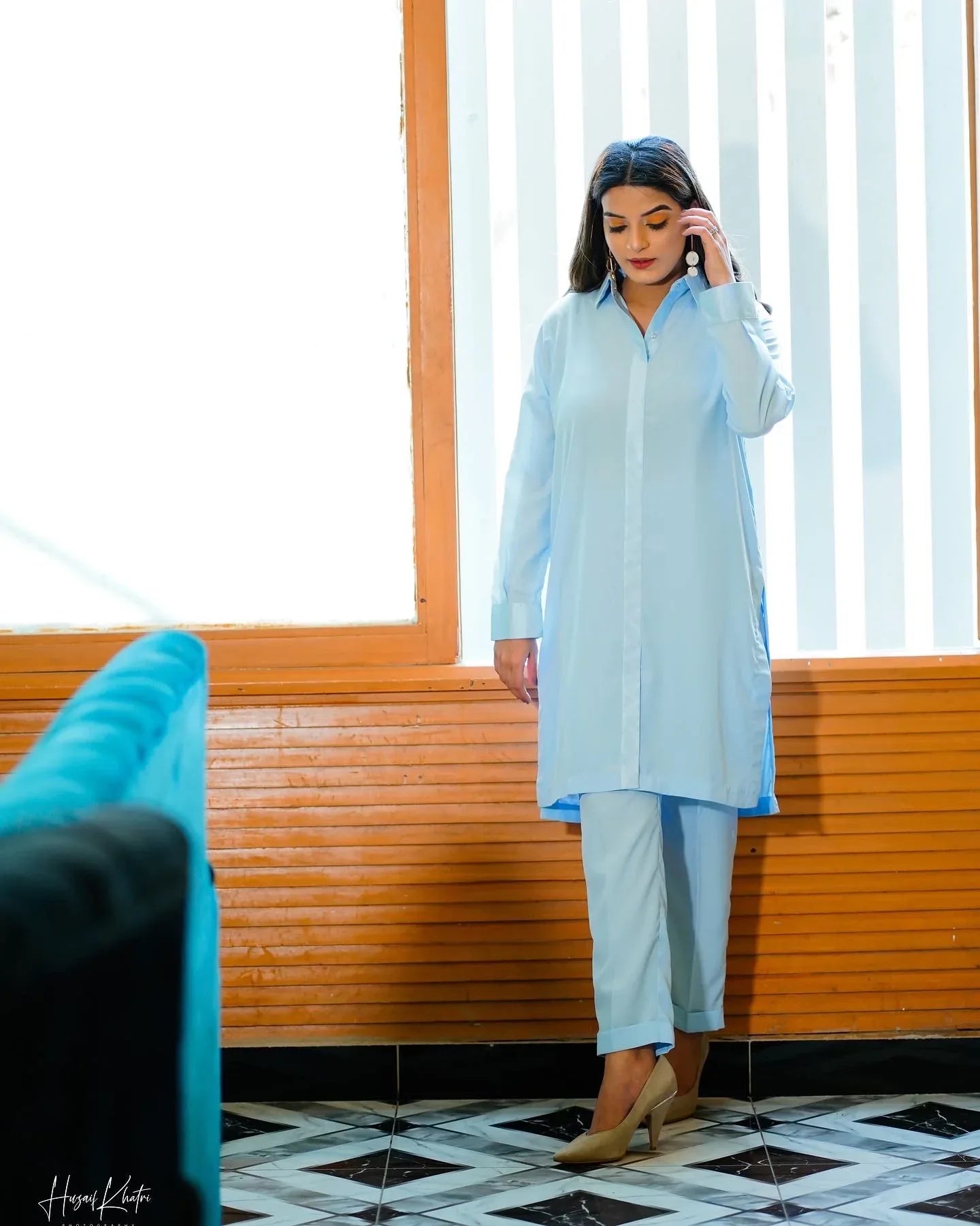 A woman in a sky blue traditional outfit poses confidently indoors. She stands near a window 