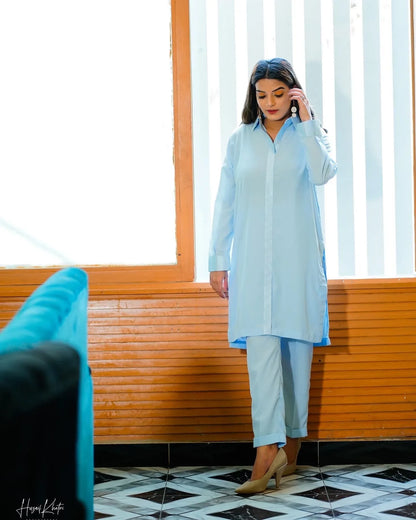 A woman in a sky blue traditional outfit poses confidently indoors. She stands near a window 