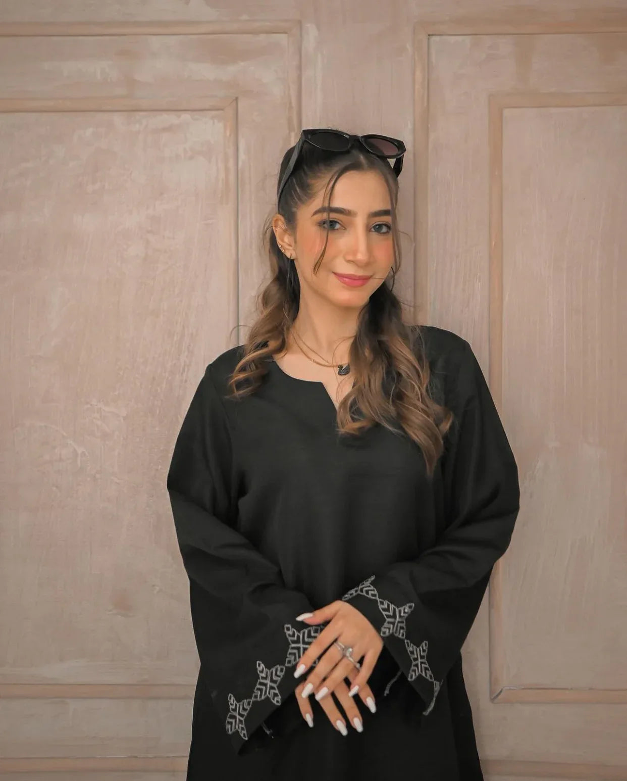 A woman in a black traditional stitched dress with white embroidery details on sleeves poses confidently in a minimalist beige room.