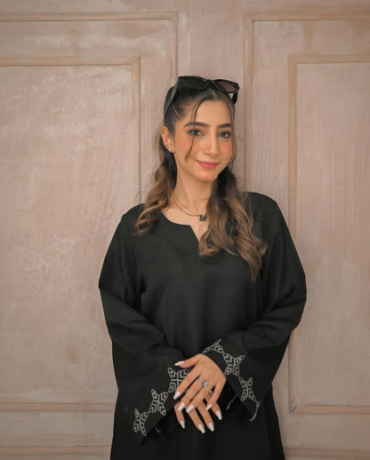 A woman in a black traditional stitched dress with white embroidery details on sleeves poses confidently in a minimalist beige room.