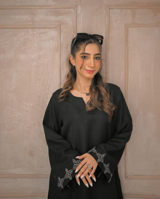 A woman in a black traditional outfit with white embroidery details on sleeves poses confidently in a minimalist beige room.