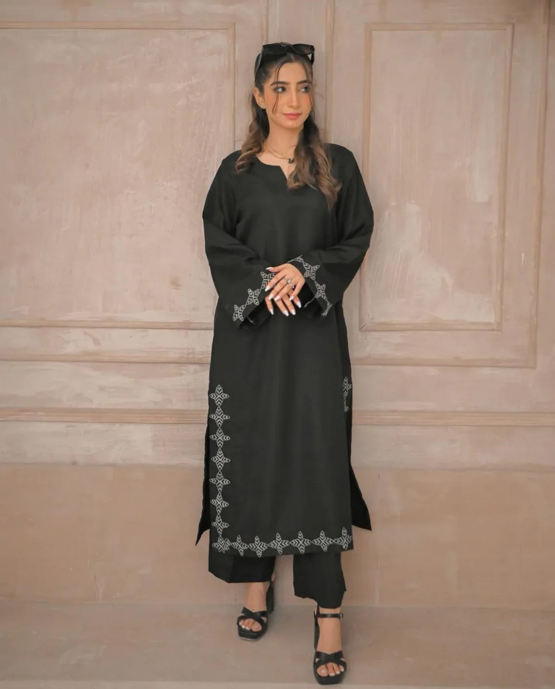 A woman in a black stitched dress with white embroidery details on sleeves and daman, poses confidently in a minimalist beige room.