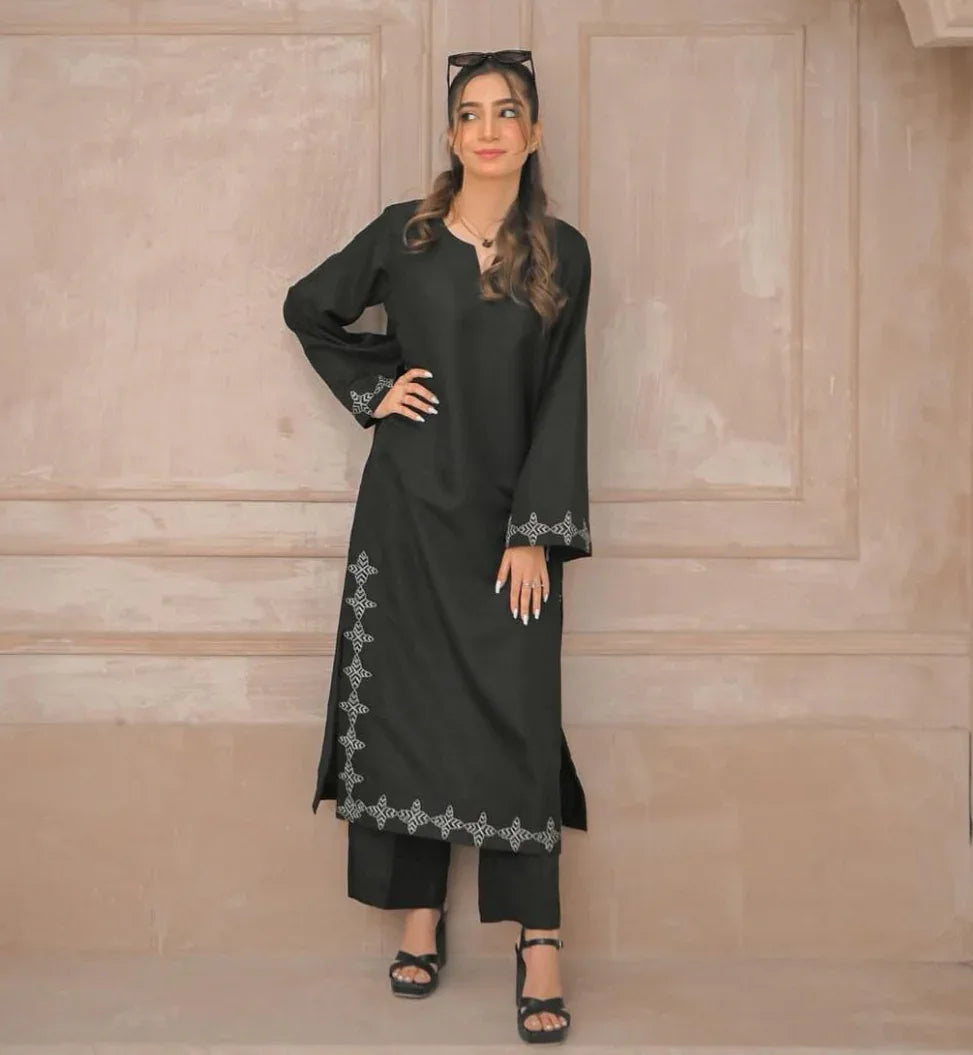 A woman in a black stitched dress with white embroidery details on sleeves and daman, poses confidently in a minimalist beige room.