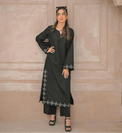 A woman in a black stitched dress with white embroidery details on sleeves and daman, poses confidently in a minimalist beige room.
