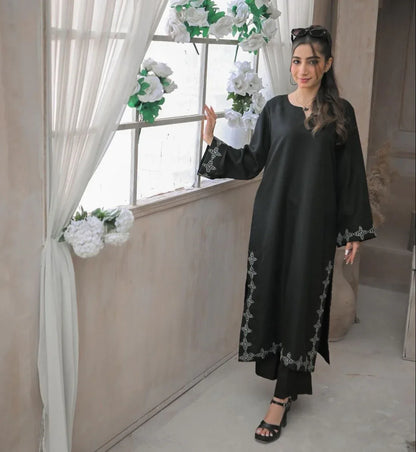 A woman in a black stitched dress with white embroidery details on daman and sleeves, poses confidently beside a window in a minimalist beige room.