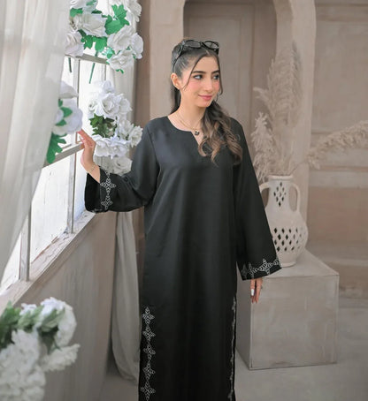 A woman in a black stitched dress with white embroidery details on sleeves and daman, poses confidently beside a window in a minimalist beige room.
