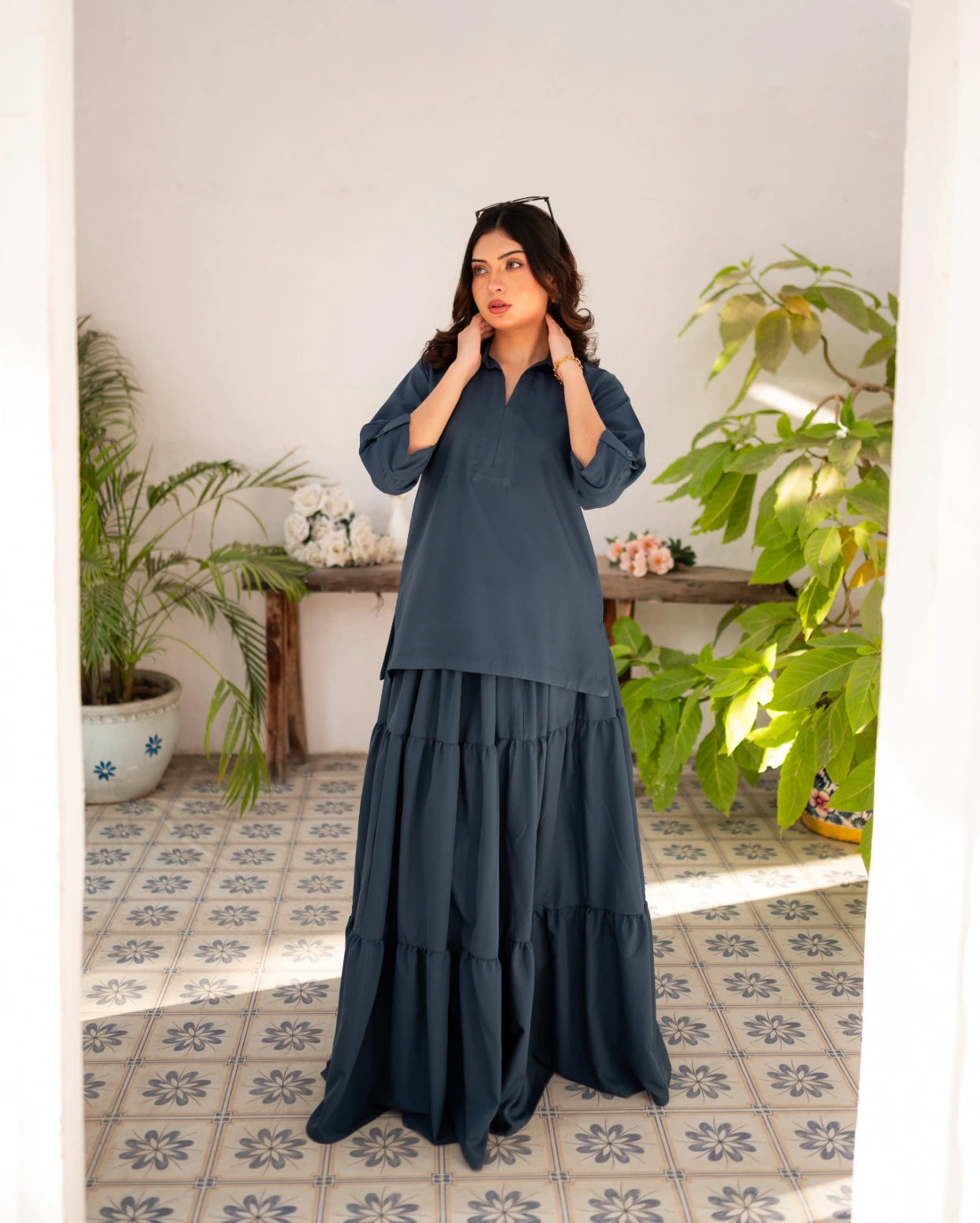 Woman wearing a steel blue skirt and shirt in a room with plants and a tiled floor.