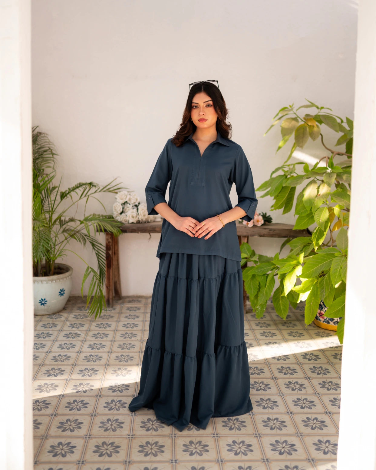 Woman wearing a steel blue skirt and shirt standing in a room with plants and a wooden table.