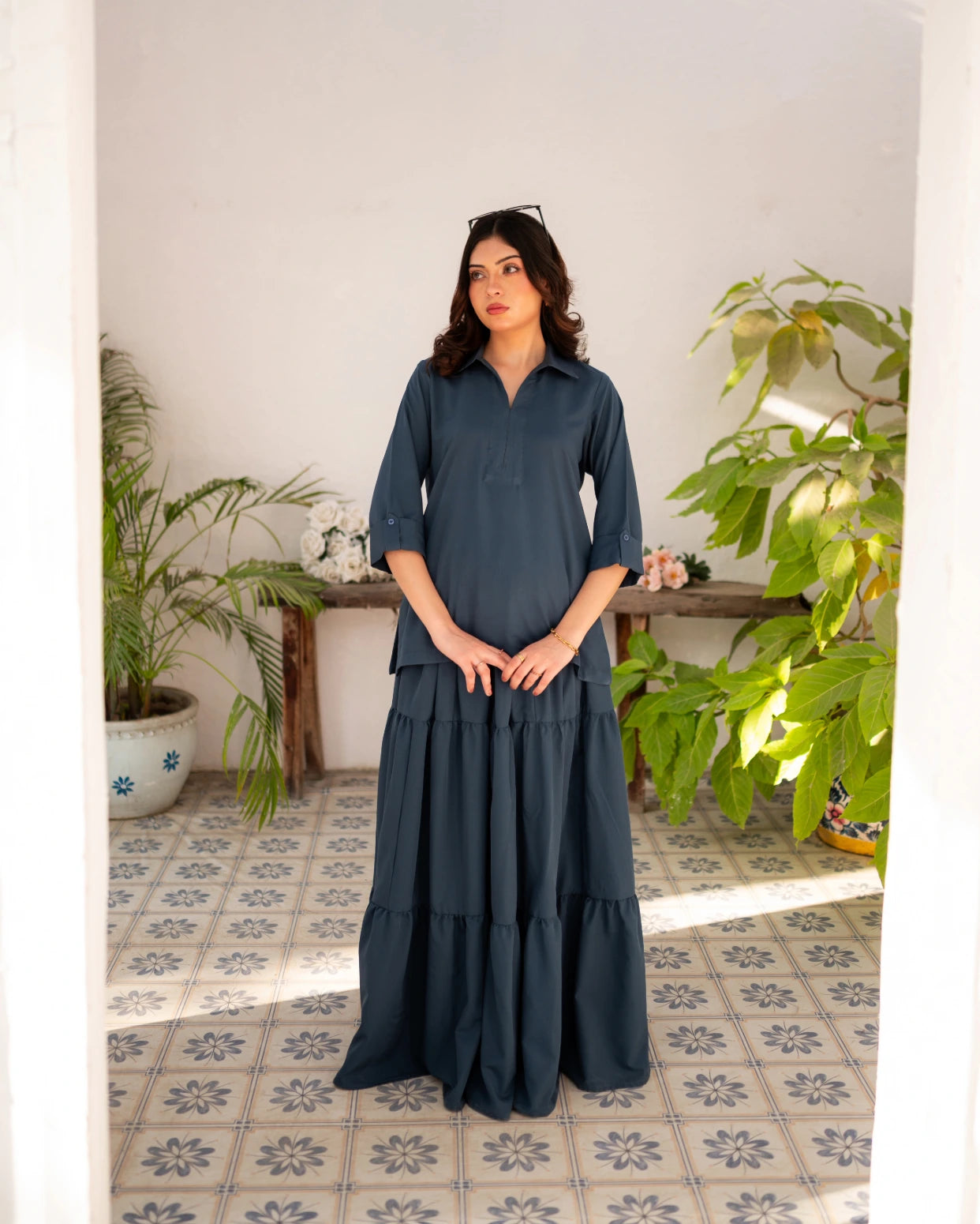 Woman in a steel blue skirt and shirt standing in a room with plants and a tiled floor.
