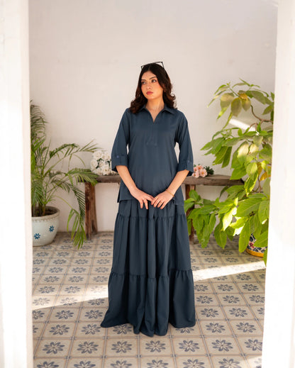 Woman in a steel blue skirt and shirt standing in a room with plants and a tiled floor.